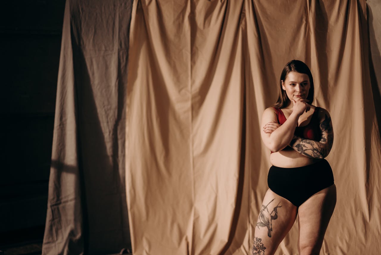 Fashion portrait of a confident woman posing in underwear with tattoos, set against a draped fabric backdrop.